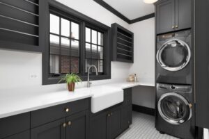 Renovated laundry room with dark cabinets and a farmhouse sink. A window looking out from over the sink lets in natural light, and the stacked charcoal gray washer and dryer bring the room together. This is a great way to maximize your budget while doing a remodel.